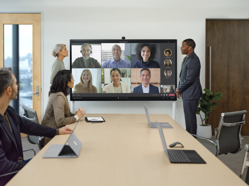 Dva kolegové prezentují na zařízení Surface Hub 2 S během konferenčního hovoru v Teams s fyzicky přítomnými i vzdálenými členy týmu.