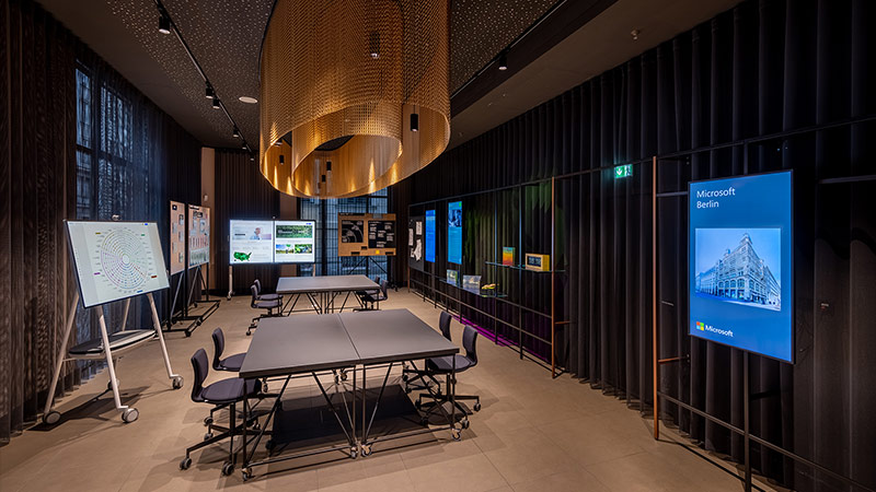 Cloud & AI Center: Das Foto zeigt einen großen Raum mit dunklen Vorhängen an allen Wänden. In der Mitte steht ein Tisch mit Stühlen, darüber ist ein Kronleuchter installiert. Der Tisch wird von Bildschirmen umrandet, auf denen man Beispiele für den Einsatz von künstlicher Intelligenz sieht.