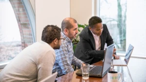 Drei Männer sitzen zusammen an einem Holztisch und sehen sich etwas auf ihren Laptops an