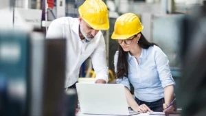 Eine Frau und ein Mann mit Schutzhelmen arbeiten in einer Werkshalle an einem Laptop