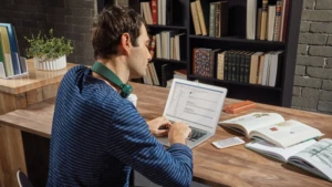 Ein Mann sitzt mit seinem Laptop an einem Schreibtisch. Neben dem Computer liegen einige aufgeschlagene Bücher.