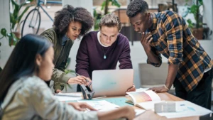 Auf dem Bild sind 4 Studenten zu sehen, die miteinander reden und am Computer arbeiten.