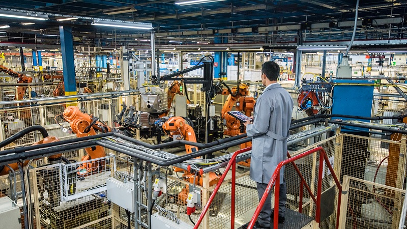 Ein Mann im grauen Kittel arbeitet an einem Tablet in einer großen Fabrikhalle