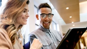 Eine Studentin und ein Student im Campusgebäude, lächeln und schauen zusammen auf das Tablet (Bildschirm nicht gezeigt).