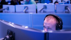 Das Bild zeigt einen Mitarbeiter mit Kopfhörern auf einen Bildschirm blickend. Im Hintergrund befinden sich weitere Mitarbeiter vor Computern.