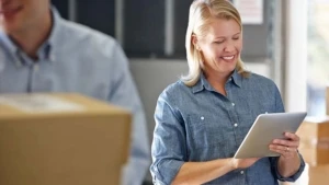 Eine Frau, umgeben von Paketen, blickt auf einen Tablet Computer.