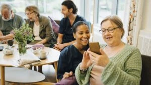 Eine ältere Dame in einer Altenpflegeeinrichtung schaut auf ihr Handy. Neben ihr kniet eine Altenpflegerin und schaut ihr dabei zu.