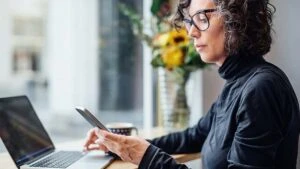 Frau sitzt mit Laptop an ihrem Schreibtisch und blickt auf ihr Smartphone