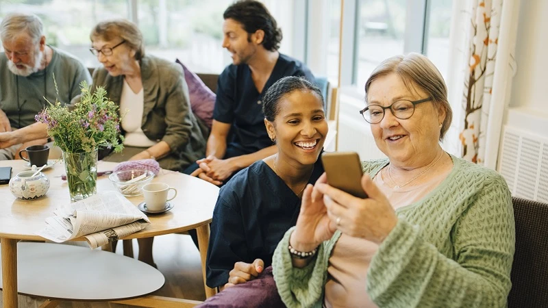 Eine ältere Dame in einer Altenpflegeeinrichtung schaut auf ihr Handy. Neben ihr kniet eine Altenpflegerin und schaut ihr dabei zu