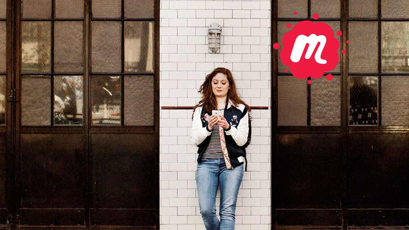 Eine Frau steht zwischen zwei Fensterfronten an eine weiße Ziegelmauer gelehnt und schaut auf ein Handy. Über ihr befindet sich das Meetup Logo. Ein kleines m in einem roten Farbklecks. 