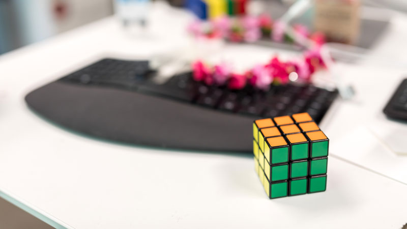 Nahaufnahme von Rubiks Würfel auf weißem Schreibtisch. Schwarze Tastatur, rosa Blumenketten und andere unscharfe Elemente, die im Hintergrund sichtbar sind.