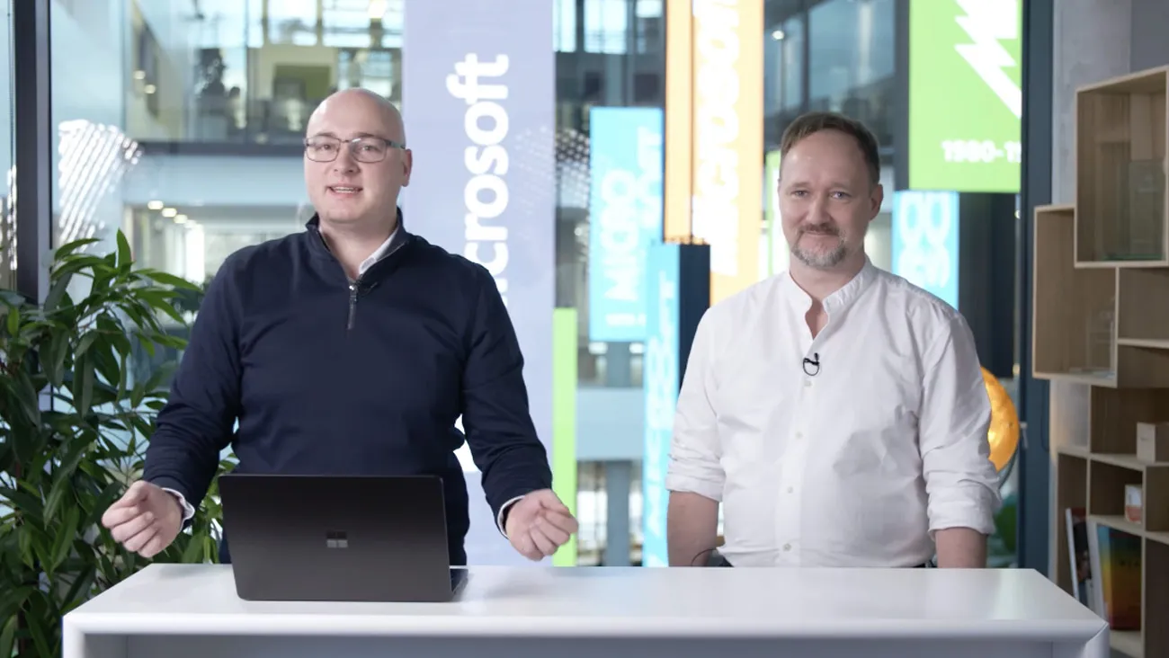 Zwei Männer stehen als Moderatoren an einem Tisch, vor Ihnen Laptops.