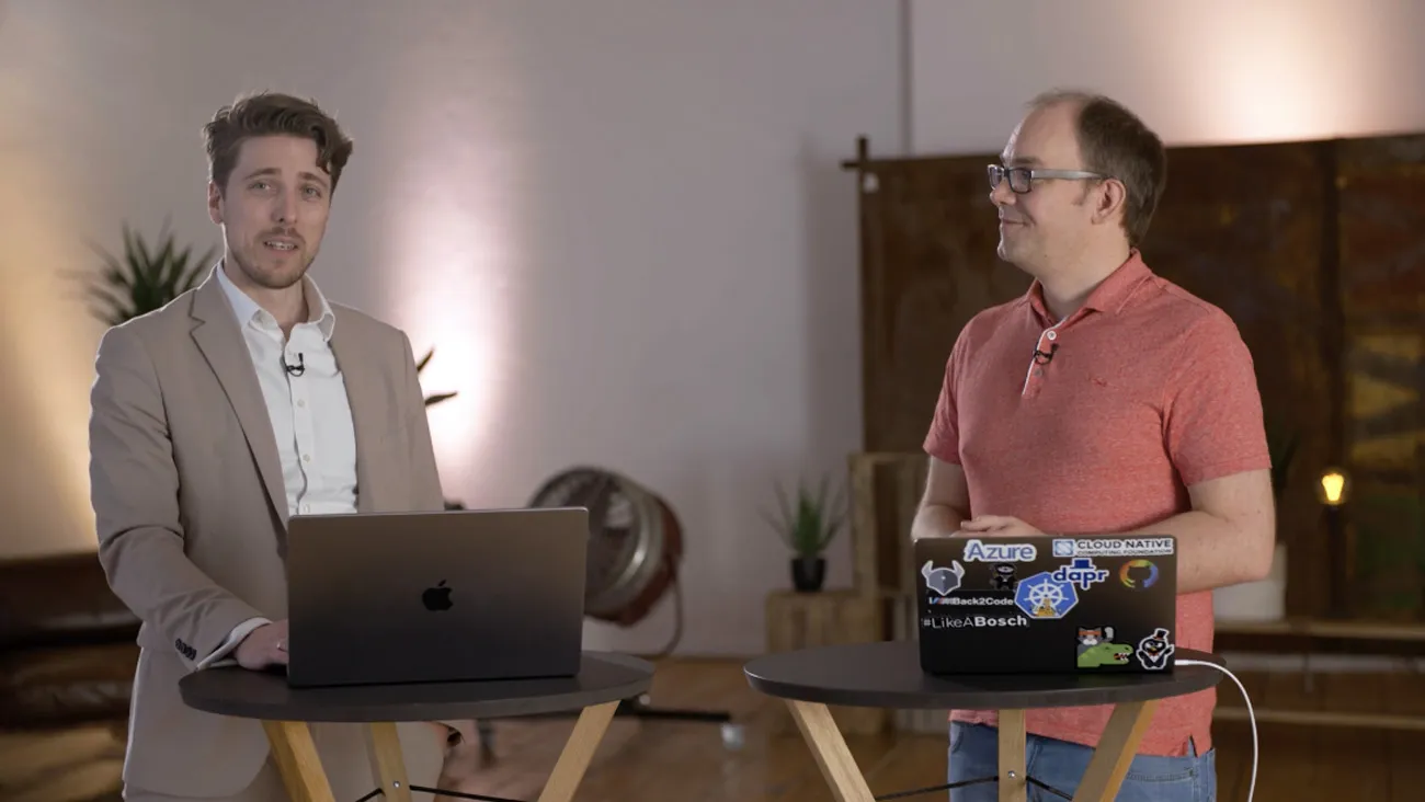 Zwei Männer stehen als Moderatoren an einem Tisch, vor Ihnen Laptops.