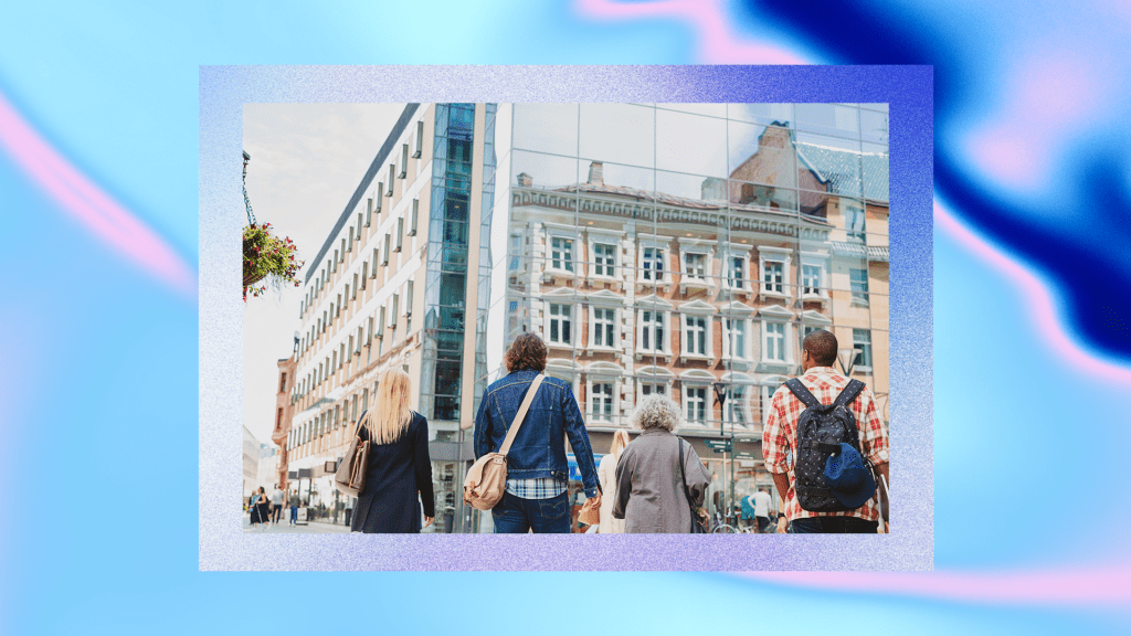 Four people walking towards a modern glass building that reflects the image of an older, ornate building with multiple windows and decorative architectural details.