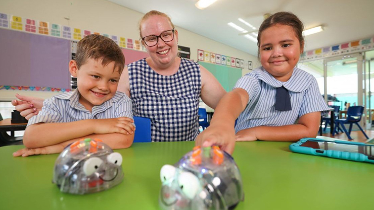 A woman and two children smile at the camera while playing with small robots on a green table