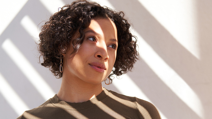 A woman with curly hair and earrings looks to the right, with sunlight casting striped shadows across her face and body.