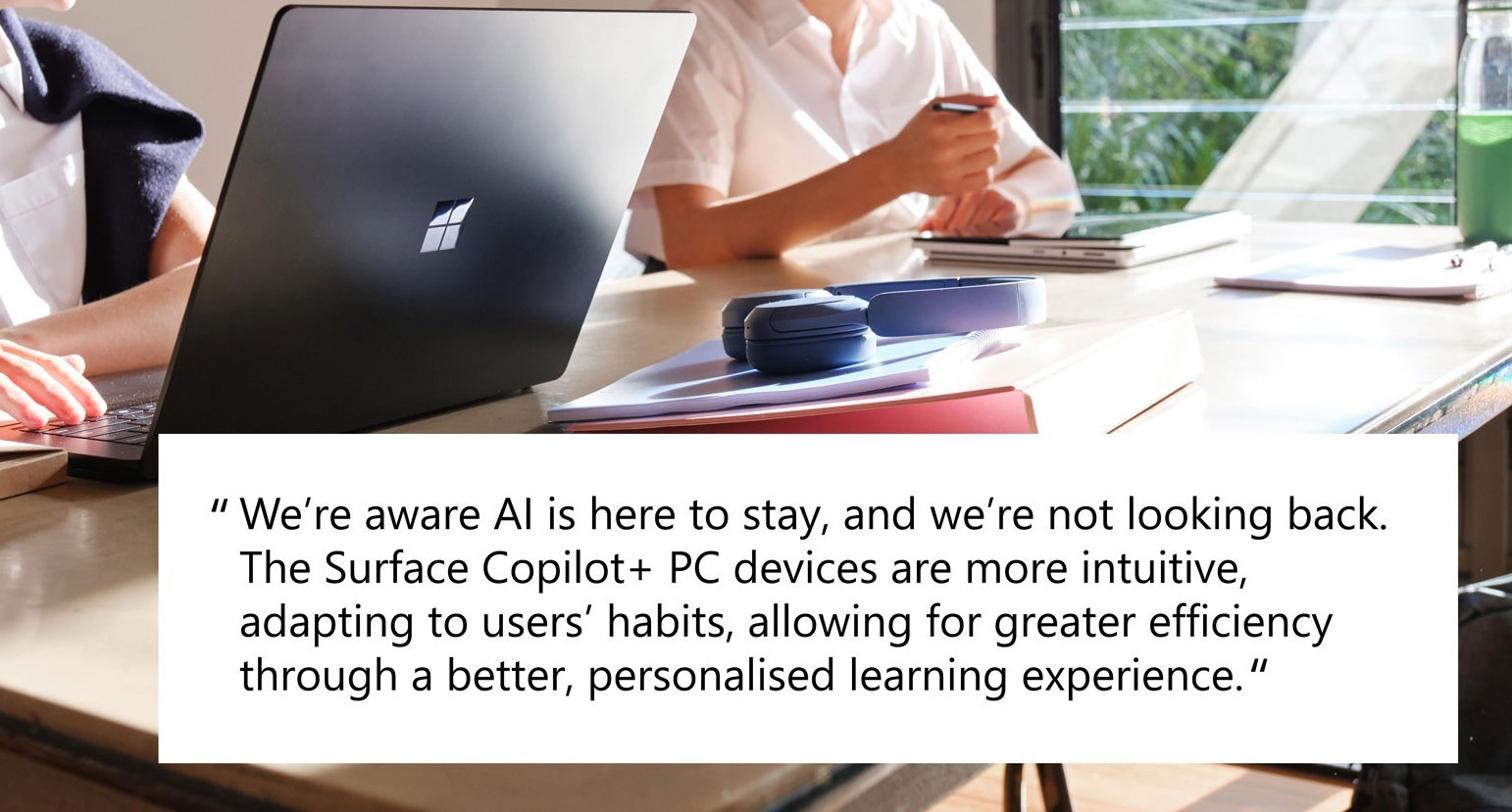 A person works on a black Microsoft Surface laptop, with another person visible in the background, while a quote about AI and Surface Copilot+ PC devices is displayed at the bottom. 'We're aware AI is here to stay, and we're not looking back. The Surface Copilot+ PC devices are more intuitive, adapting to users' habits, allowing for greater efficiency through a better, personalised learning experience.'