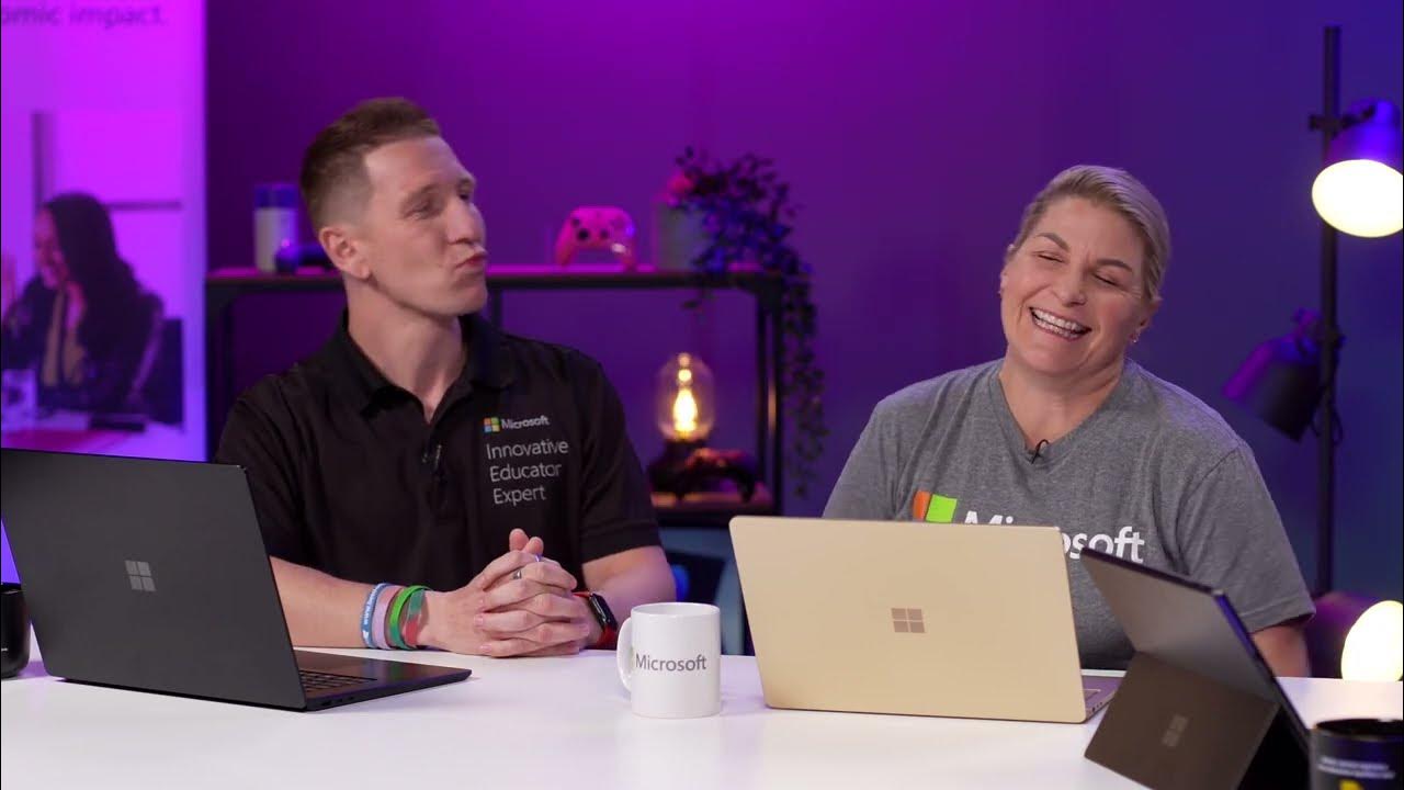 Two people are seated at a desk with Microsoft Surface laptops in front of them, with a play button overlay at the center indicating that this is a video.