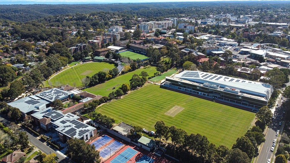 An aerial view shows a sprawling campus with multiple green sports fields, tennis courts, and buildings with solar panels, surrounded by a suburban area with many trees and houses.