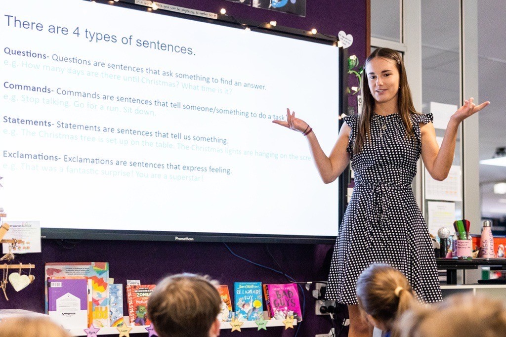 A teacher in a polka dot dress explains sentence types to students in a classroom.