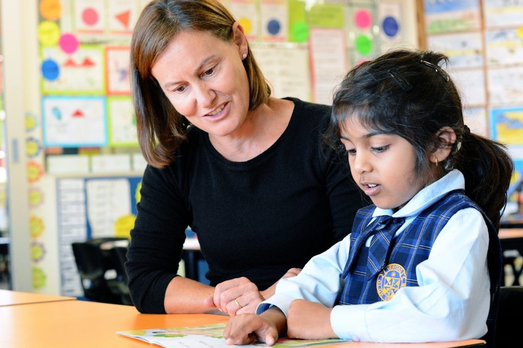 A female teacher helps a young girl in a school uniform read from a book at a desk.