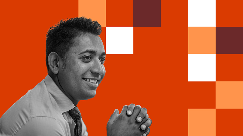 A smiling man wearing a shirt and tie with hands clasped, against a red-orange and white checkered background.
