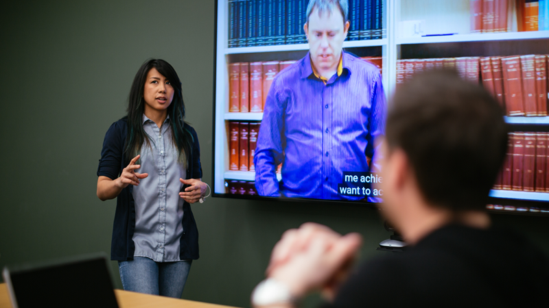 1. Photograph of Erika Martinez, a woman who is deaf, presenting using a screen with captions in a conference room.