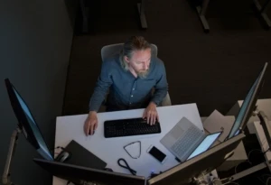 Top-down view of a bearded man in a gray/blue shirt seated at a desk working on a Surface laptop connected to three large monitors.