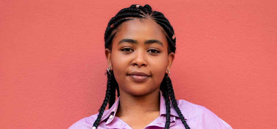 A woman with braided hair stands in front of a pink wall, smiling at the camera