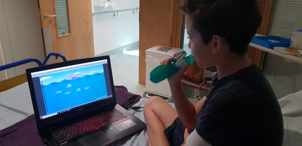 Young boy breathes into a device and plays a game on a monitor in a hospital room