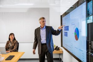 Male businessman in suit giving presentation in office conference room. He is pointing at a large monitor screen, which displays Dynamics 365