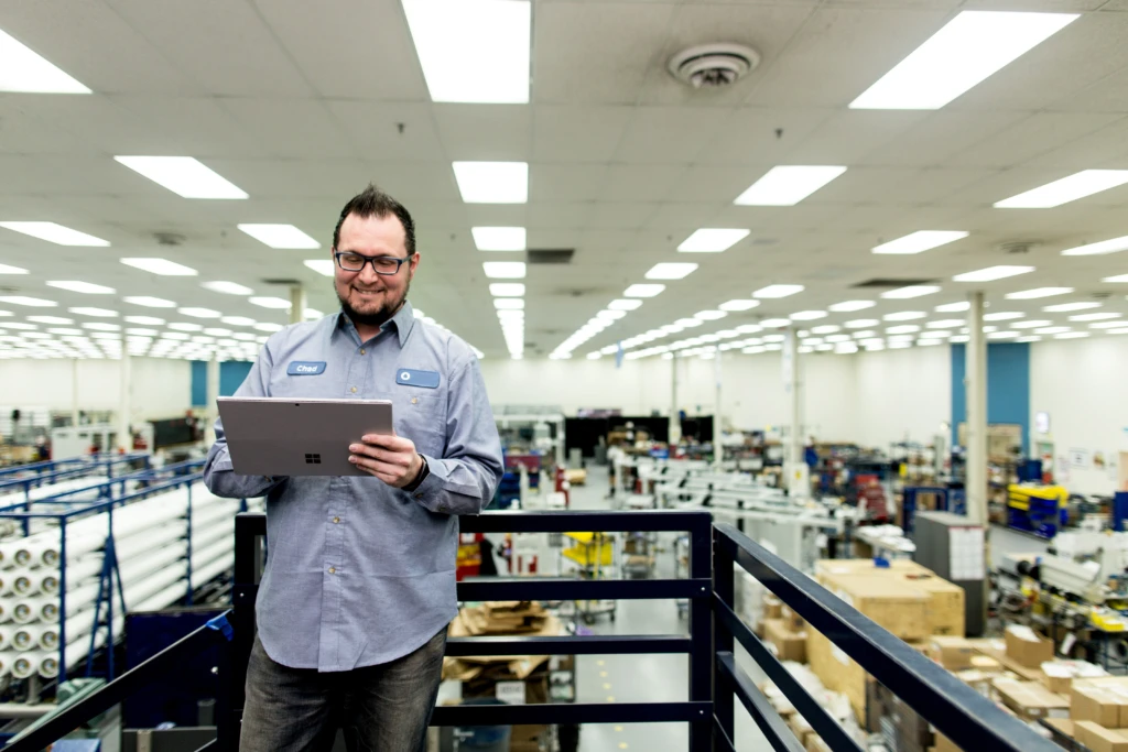 Male factory worker standing above manufacturing plant floor, smiling and looking down at Surface Pro