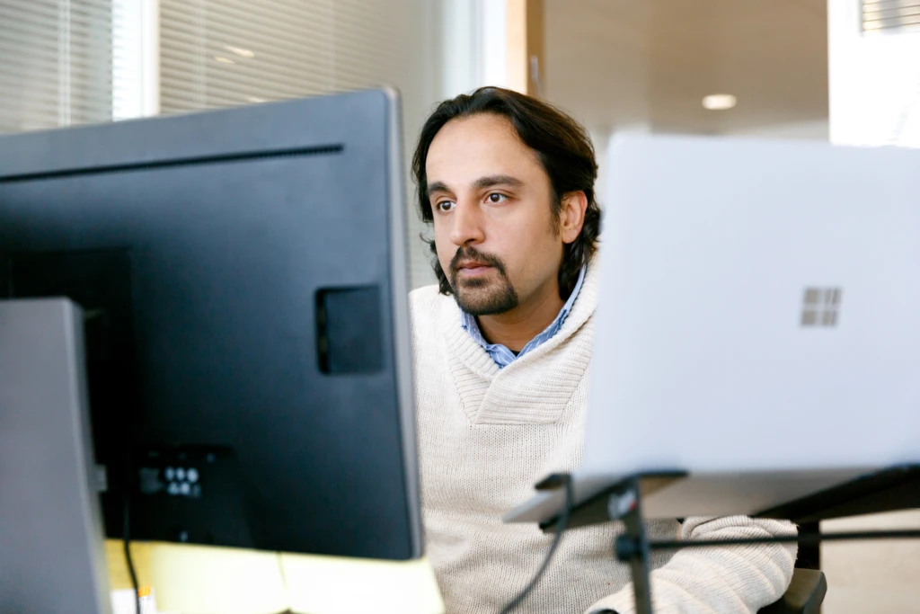 Male office worker looking at monitor on desk (screen not shown). A open Surface Laptop is also on his desk (screen not shown).