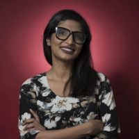 Picture of a smiling woman in a floral shirt with long dark hair and glasses. She is standing with her arms crossed in front of a red background.
