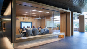 A group of employees sit around a table, working at their laptops during a meeting in a glass-walled office with large screen.