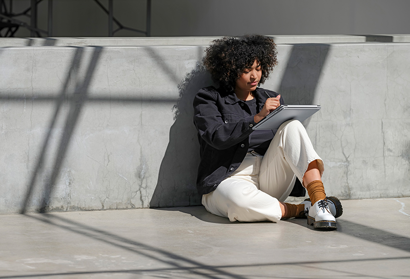 A young woman is sitting outside on the ground and leaning against a wall, working on a laptop