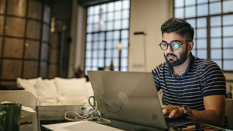 A man working on a laptop at a glass table in a modern, softly lit room with large windows and a couch in the background. The scene includes a green mug, earbuds, and papers on the table, suggesting a focused work or study environment.