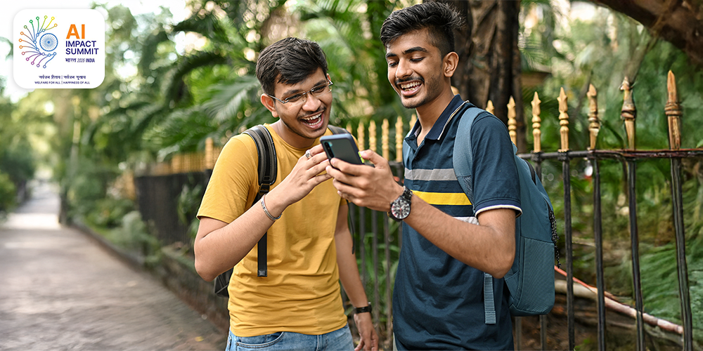 Two young men standing outdoors near a metal fence with greenery in the background, both wearing backpacks and looking at a smartphone held by one of them. casual interaction or sharing information, with one person in a yellow shirt and the other in a navy blue shirt with stripes | AI IMPACT SUMMIT भारत 2026 INDLA सर्वजन रिवाद/ सर्वजन सुखाद WELFARE FOR ALL | HAPPINESS OF ALL