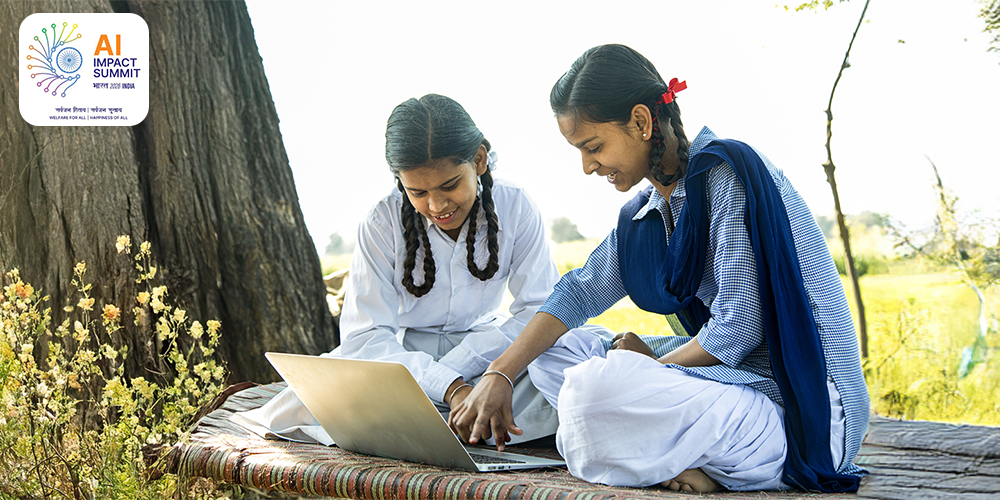 Two girls sitting outdoors on a woven cot using a laptop, with one girl pointing at the screen. The setting includes natural background with trees and grass, highlighting a rural or semi-rural learning environment | AI IMPACT SUMMIT भारत 2026 INDLA सर्वजन रिवाद/ सर्वजन सुखाद WELFARE FOR ALL | HAPPINESS OF ALL