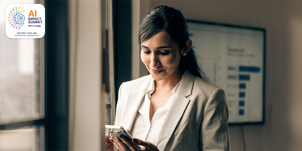 A professional woman in a beige blazer standing near a window while using a smartphone, with a blurred presentation slide visible on a screen in the background. The setting suggests a business or office environment focused on communication or presentation activities | AI IMPACT SUMMIT भारत 2026 INDLA सर्वजन रिवाद/ सर्वजन सुखाद WELFARE FOR ALL | HAPPINESS OF ALL