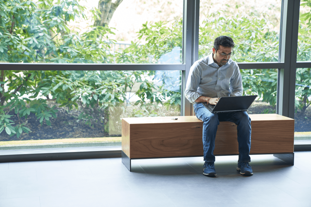 Man sitting on an indoor bench in front of windows working on a laptop