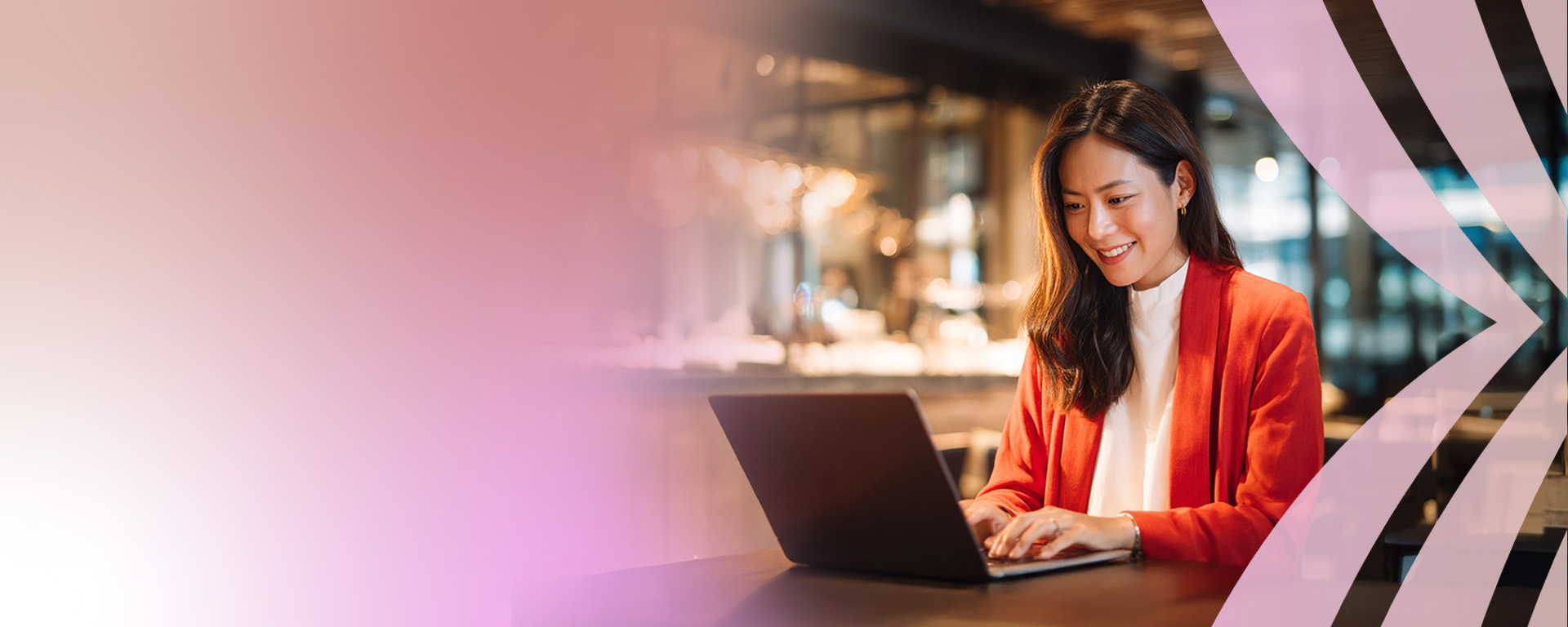 A young woman in a red blazer smiles while typing on her laptop at a table