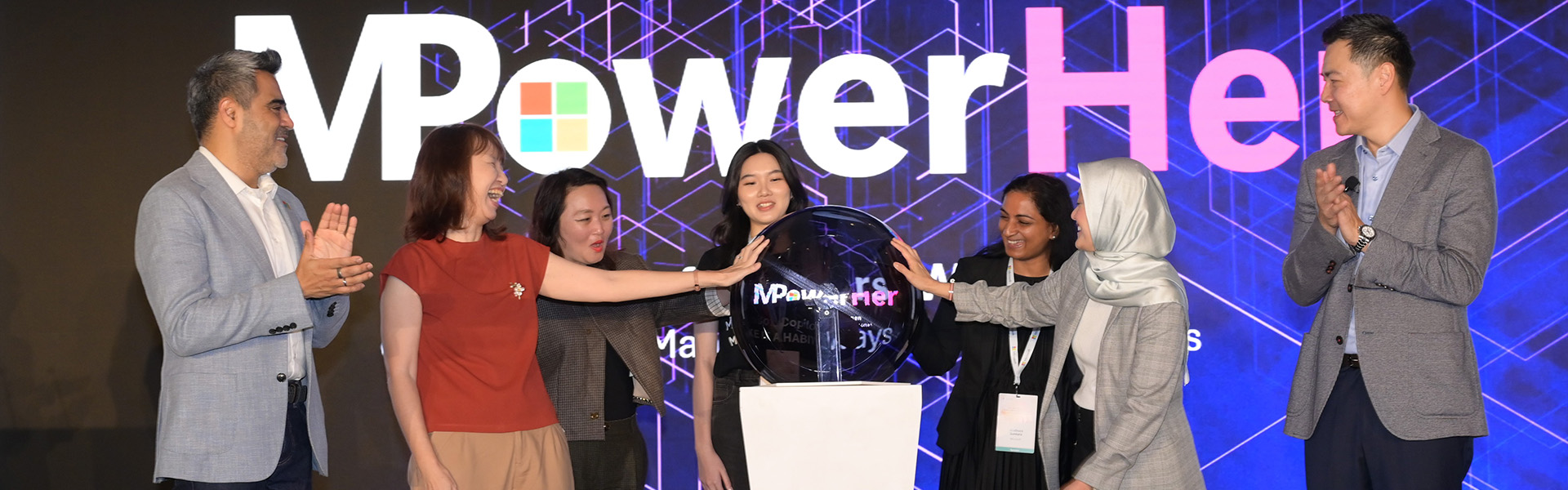 Group of speakers on a stage jointly unveiling a display at a MPowerHer event, with a branded backdrop behind them.