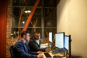 Photo of two male developers working and collaborating in an casual enterprise office. They are using Visual Studio with a multi-monitor set up.