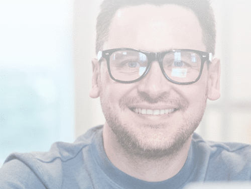 A smiling man wearing black-rimmed glasses and a blue T-shirt looks straight into the camera.
