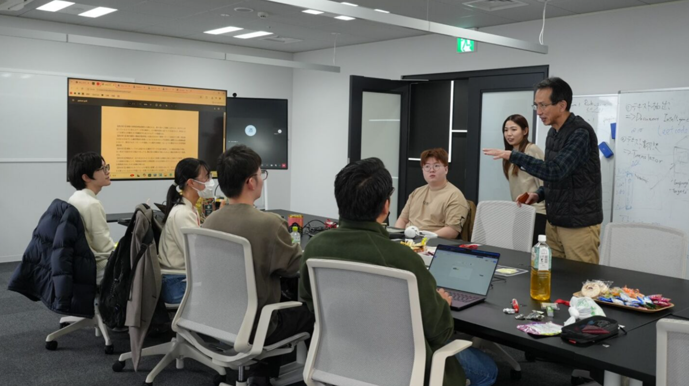 A business meeting in progress with a man standing and gesturing, addressing a group seated around a table with laptops, a water bottle, and snacks. A large screen displays text, and a whiteboard shows 'Artificial Intelligence', 'AI', and Japanese writing.
