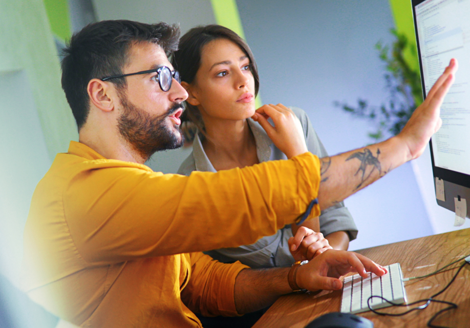 A man in a yellow shirt and glasses points at a computer screen while a woman beside him looks on intently, both collaborating in a modern office setting.