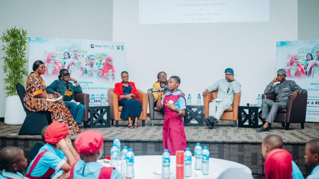 A child speaks at a panel event with six seated panelists, banners, and an audience present.