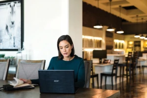 Female first line worker sitting at table in commercial retail store using Acer laptop. She is flipping through a book of fabric sample swatches, while looking at laptop.