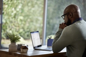 Male working remotely from his home office on a Dell Latitude 13 device. This image is available with two different screen options: one showing the device running Microsoft Outlook, and another showing a Microsoft Teams conference call with 9 people on the screen.